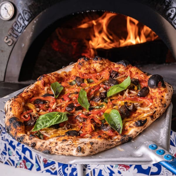 A freshly baked Doppio Zero pizza with vegetables and basil on a peel, in front of a wood-fired oven.