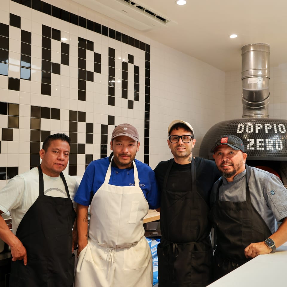 Four male chefs in aprons stand smiling in a kitchen with a large pizza oven.