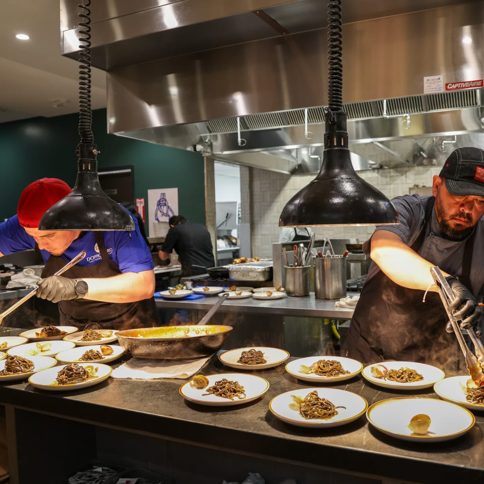 Chefs plating pasta dishes under heat lamps in a busy commercial kitchen.