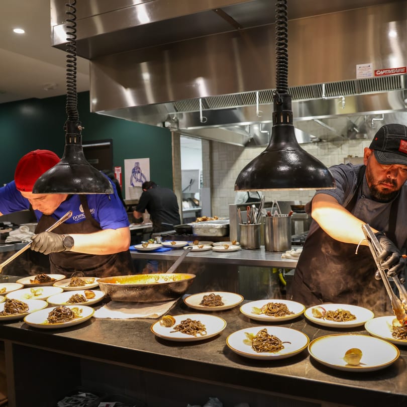 Chefs plating pasta dishes under heat lamps in a busy commercial kitchen.