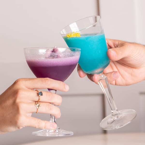 Two hands clinking glasses of vibrant purple and blue cocktails, garnished with flowers.