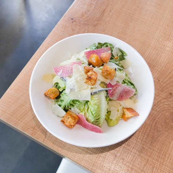 A white bowl of Caesar salad with romaine, croutons, parmesan, anchovies, and pink radish slices.
