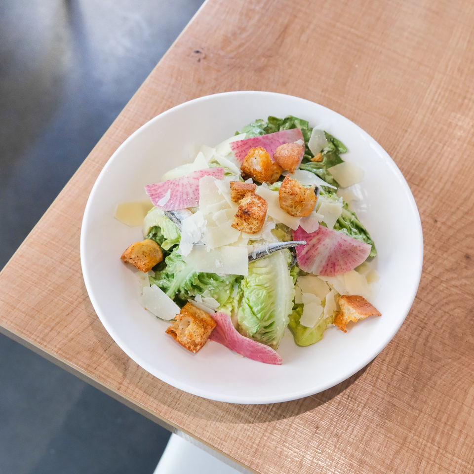 A white bowl of Caesar salad with romaine, croutons, parmesan, anchovies, and pink radish slices.