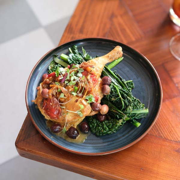 Doppio Zero's Italian chicken leg with olives, tomatoes, and greens on a dark plate.