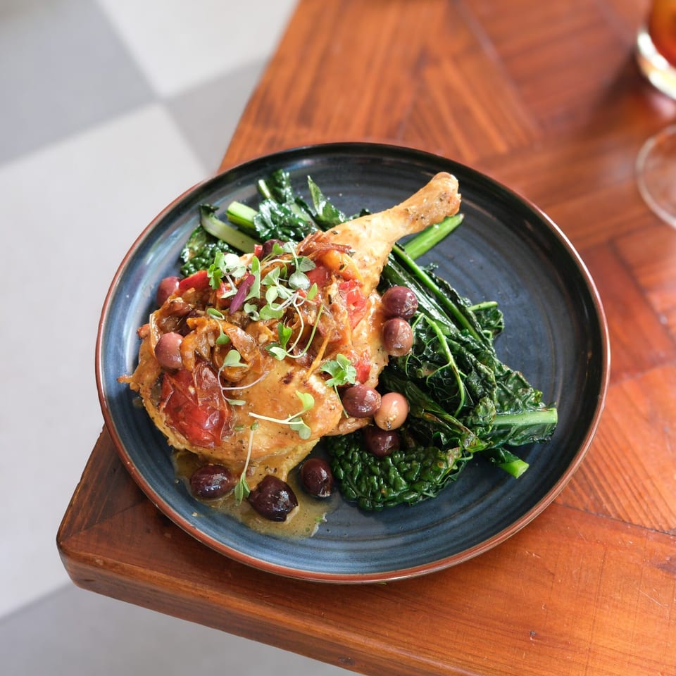 Doppio Zero's Italian chicken leg with olives, tomatoes, and greens on a dark plate.