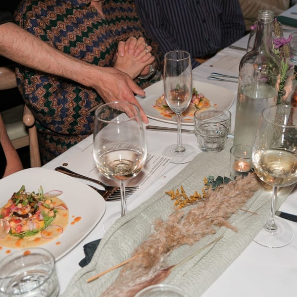 Hands clasped at a dining table with plates of food, wine glasses, and a water bottle.