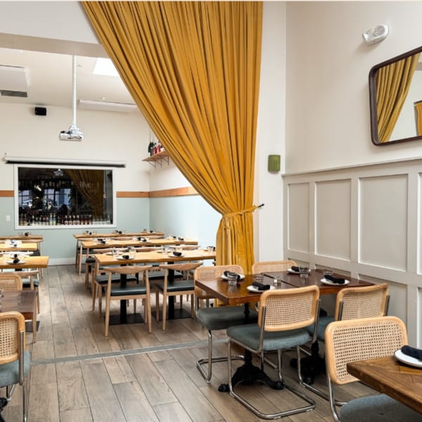 Restaurant dining room with wooden tables, cane-back chairs, and a large yellow curtain dividing the space.