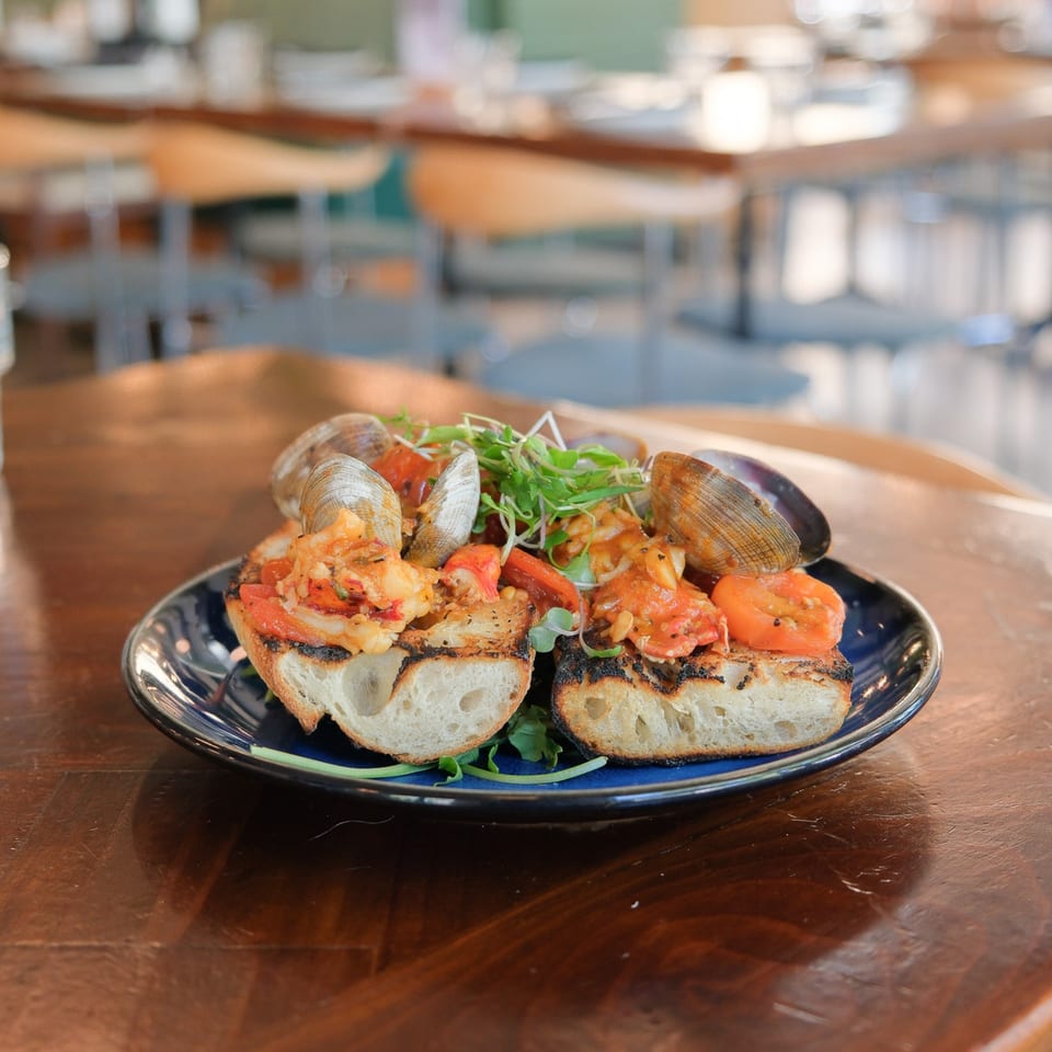 Grilled bread topped with shrimp, clams, tomatoes, and microgreens on a dark plate in a restaurant.
