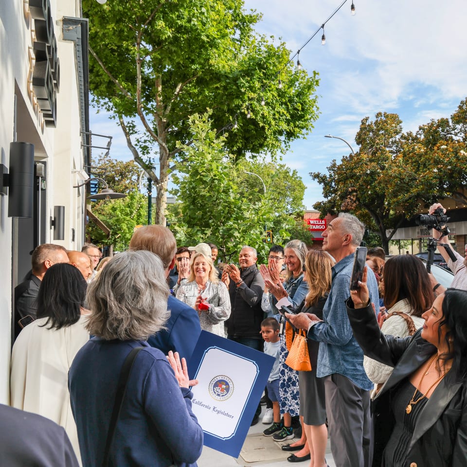 A crowd of people gathered outside a building, applauding a woman holding a framed certificate.