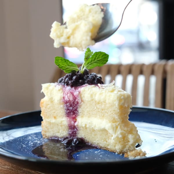 A slice of white cake with blueberry topping and mint, with a spoon taking a bite.