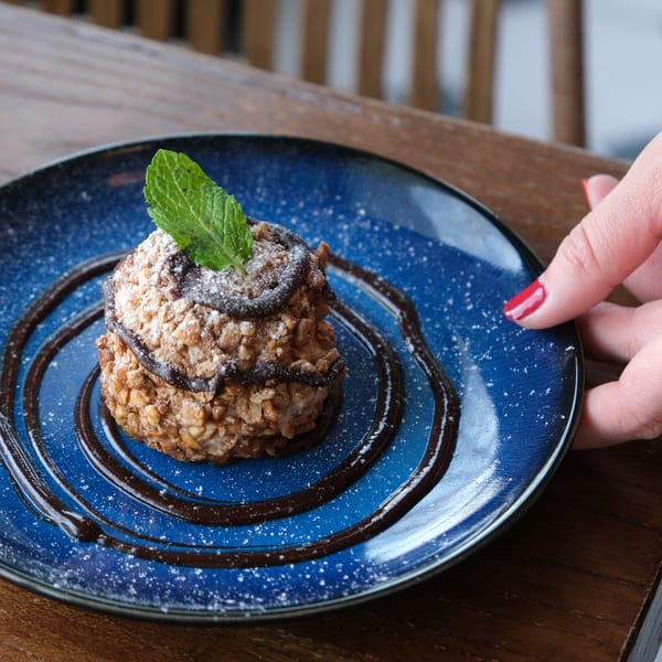 A hand with red nail polish serving a dessert with chocolate drizzle and mint on a blue plate.