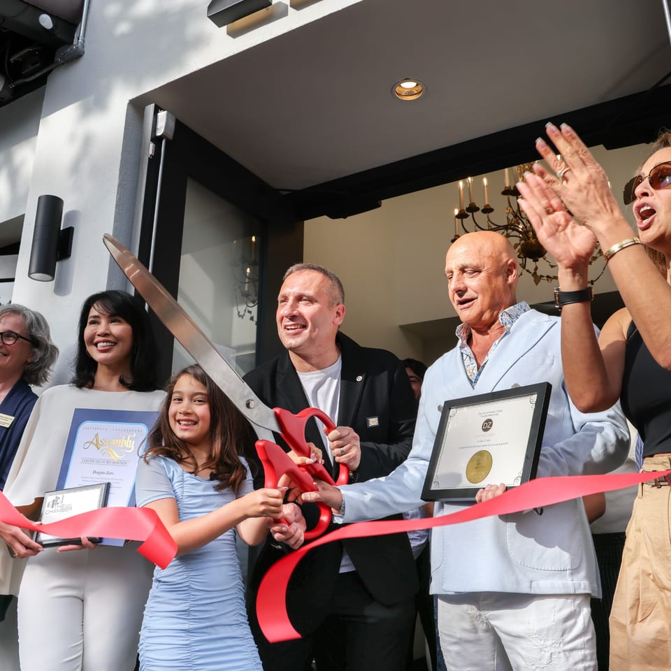 People, including a child, cutting a red ribbon with giant scissors at a grand opening event.