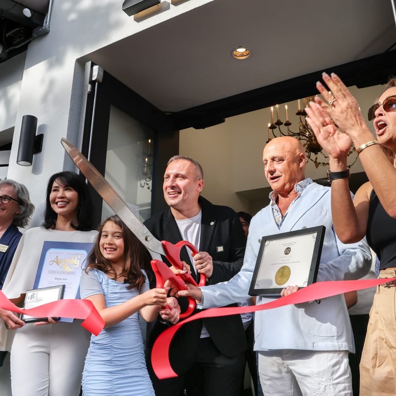 People, including a child, cutting a red ribbon with giant scissors at a grand opening event.