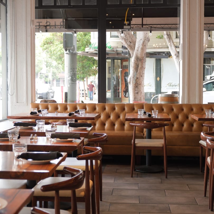 Restaurant interior with wooden tables, chairs, and a long tufted leather booth next to large windows.