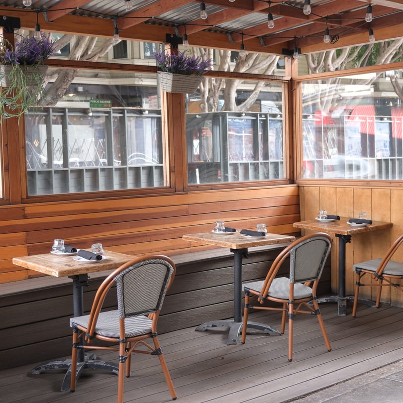 Outdoor dining patio with wooden walls, string lights, potted plants, and small tables with chairs.