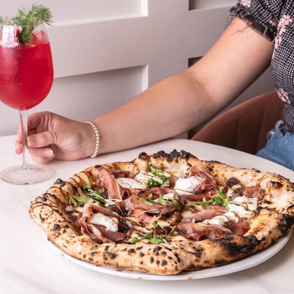 A hand holding a red cocktail next to a Doppio Zero pizza with prosciutto, burrata, and figs.