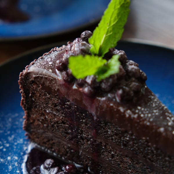 A rich slice of chocolate cake with ganache, chocolate pearls, and mint on a blue plate.