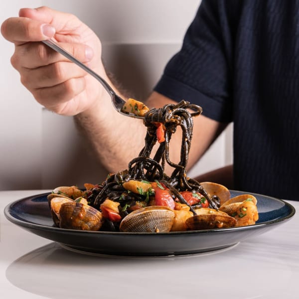 A hand twirls black squid ink pasta with clams, shrimp, and tomatoes onto a fork.