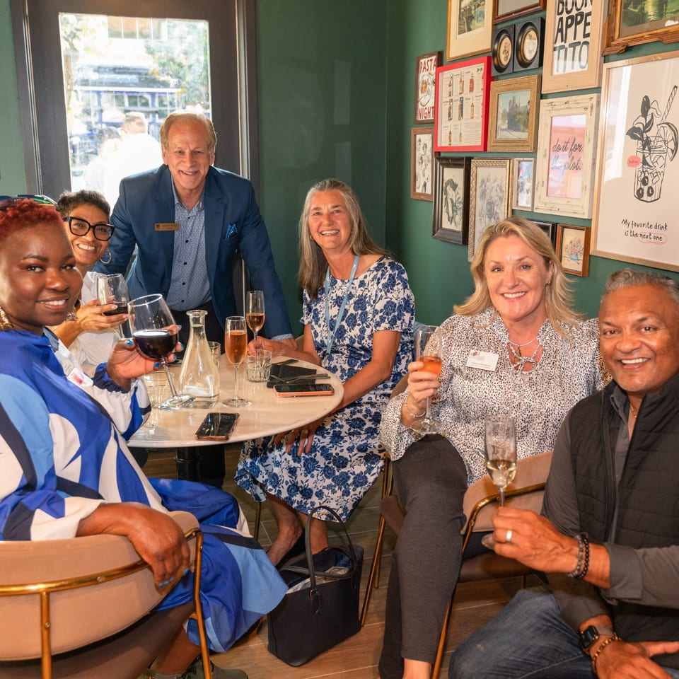 Six smiling people, holding drinks, gathered around a table in a restaurant with framed art.