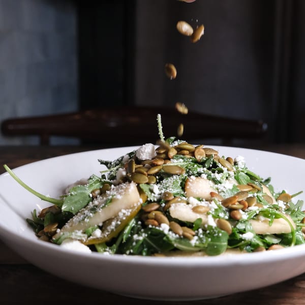 Arugula salad with pear slices, feta cheese, and pumpkin seeds being sprinkled on top.