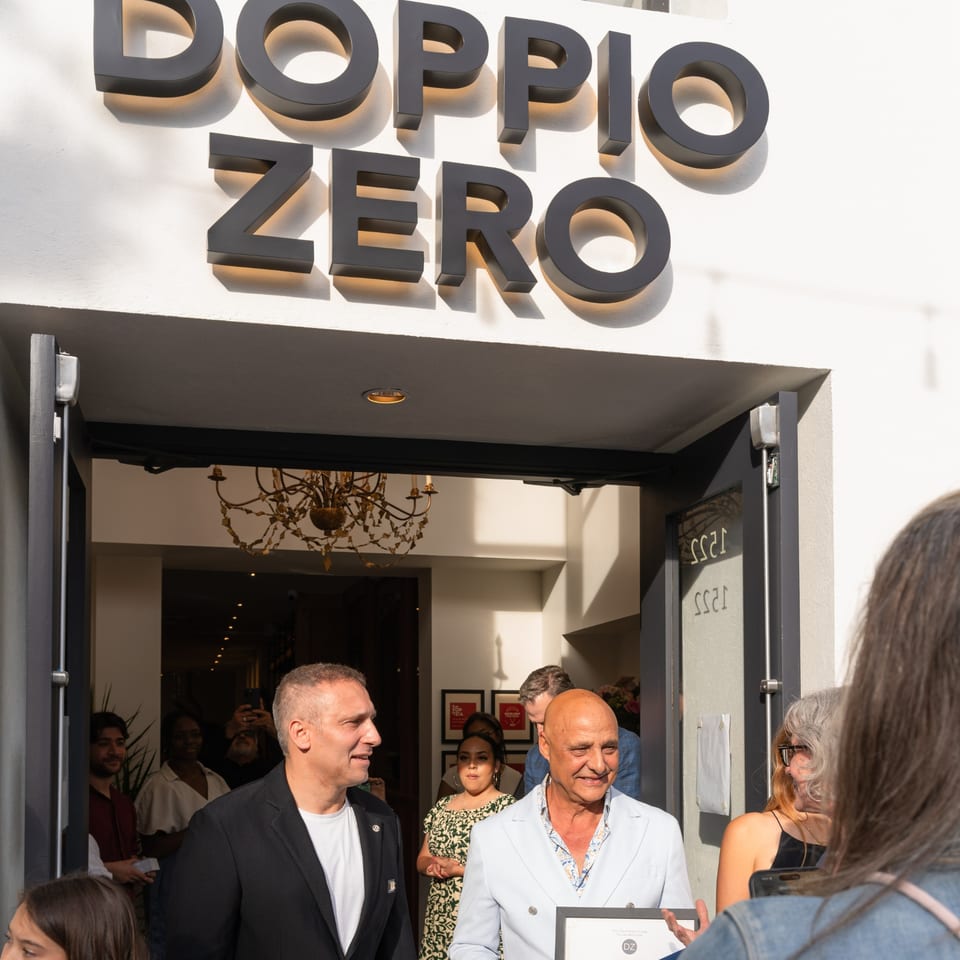 Two men smiling outside Doppio Zero restaurant, with a crowd and chandelier visible inside.