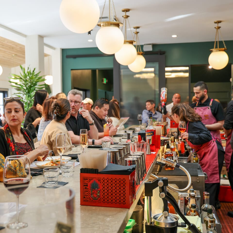 Diners and bartenders at a lively bar, with drinks and food on the counter.