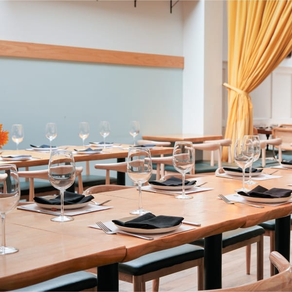 A long wooden dining table set with plates, black napkins, silverware, and wine glasses in a restaurant.