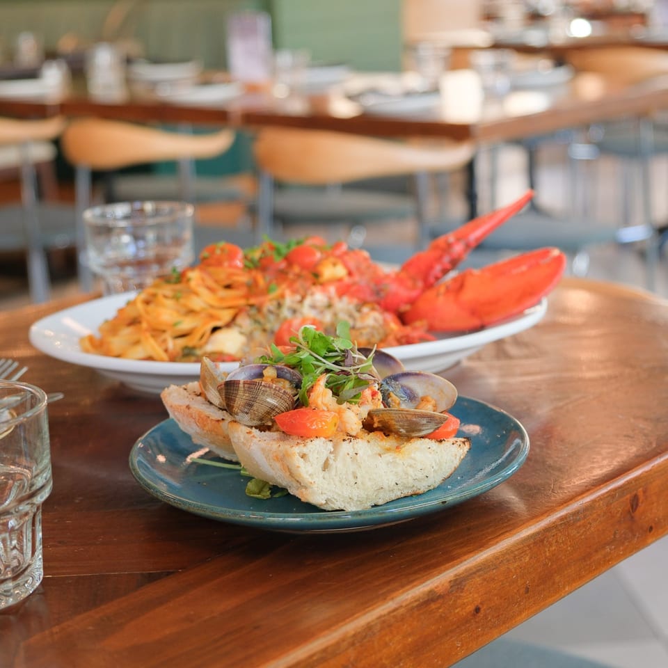 Seafood dishes with clams, shrimp, lobster, pasta, and herbs served on a wooden restaurant table.