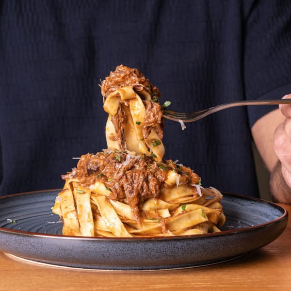 A person twirling pappardelle pasta with rich beef ragu and grated cheese onto a fork.