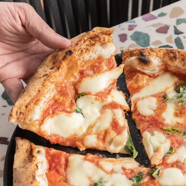 A hand lifts a slice of hot, cheesy Margherita pizza from a dark plate on a colorful table.