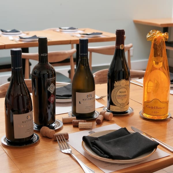 Five wine bottles, including Kettmeir, Masi, and Ca'del Bosco, on a light wooden table with corks.