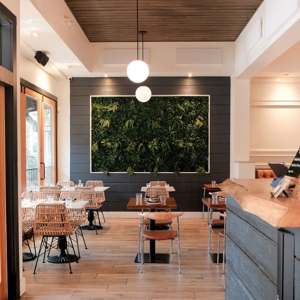 Modern restaurant interior with a large green plant wall, wooden floors, and rattan chairs.