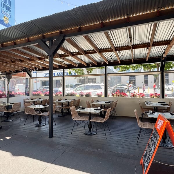 An empty outdoor patio with tables and chairs under a corrugated metal roof, facing a street.