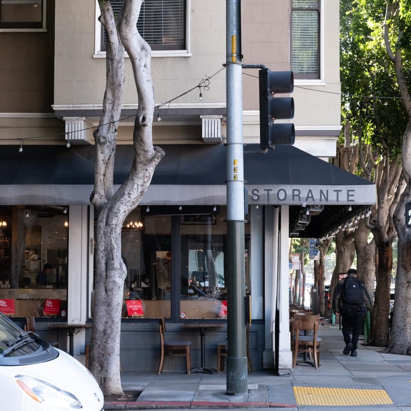 A restaurant storefront with a black awning, outdoor seating, and a person walking on the sidewalk.