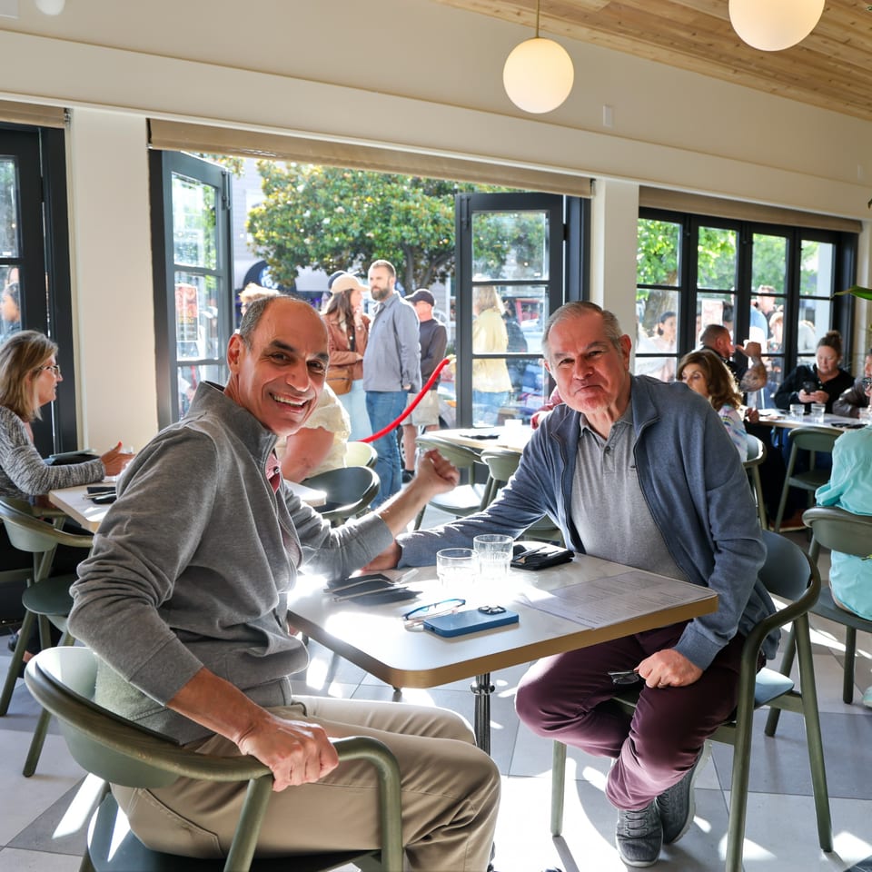 Two men smiling at a table in a bright, modern restaurant with large windows.