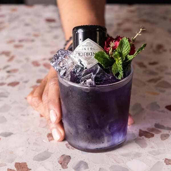 A hand holds a purple cocktail with ice, a miniature gin bottle, mint, and red flowers.