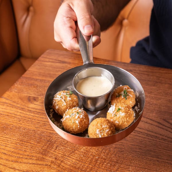 A hand holding a small pan with five golden-brown arancini balls and a dipping sauce.