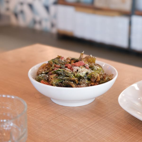 A white bowl of crispy Brussels sprouts with red peppers and grated cheese on a wooden table.