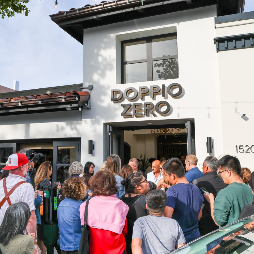 A crowd of people gathered outside the Doppio Zero restaurant entrance on a sunny day.